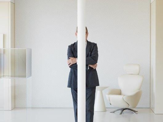 Man in suit with arms crossed standing behind a post in an all-white room. His face is not visible.