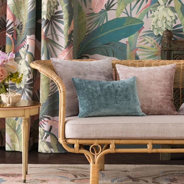 Rattan sofa with cushioned seat and accent pillows. In background wall and drapes are in tropical plant pattern.